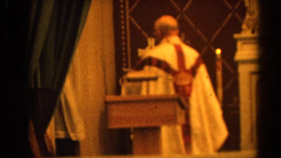 A Catholic priest in ornate vestments is seen from behind near drapery and altar furnishings in the 1964 church interior.