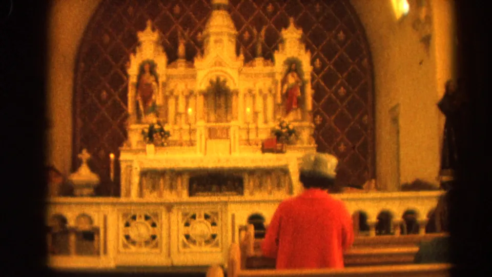 1964 Harvard, Illinois 8mm frame showing a person in a red sweater and light hat seated before an ornate church altar.