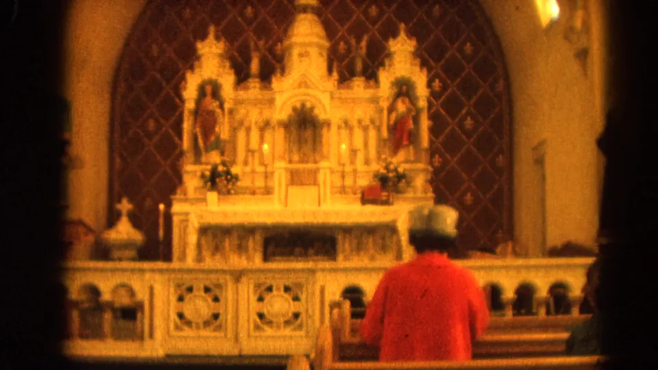 Home-movie frame from 1964 Harvard, Illinois showing a worshipper from behind and an ornate church altar in warm light.