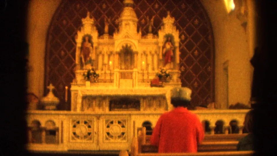1964 8mm church-interior frame from Harvard, Illinois, with a person seated before an altar, candles, and religious statues.
