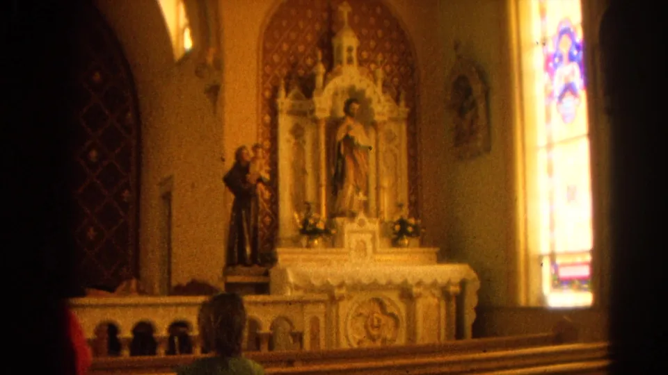 1964 home-movie frame inside a Harvard, Illinois church, showing an altar, saint statue, stained glass, pews, and a worshipper from behind.