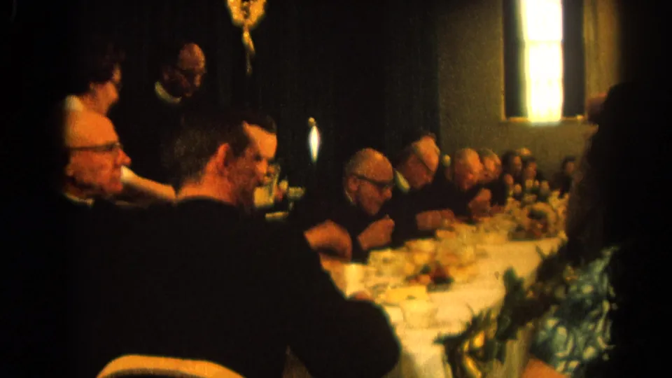 1964 Harvard, Illinois communal parish meal with clerics and other adults seated at a long table in a dim interior hall.