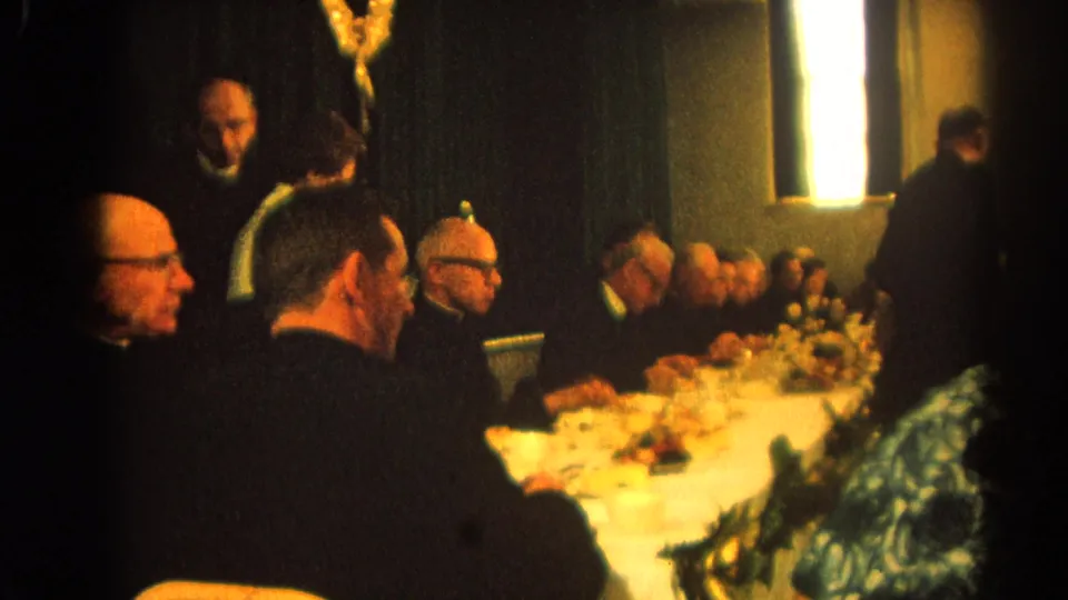 1964 Harvard, Illinois parish meal scene showing clerics and adults seated around a long table with food, flowers, and low window light.