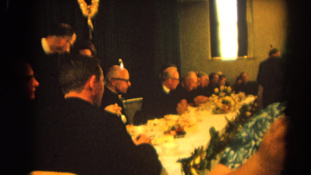Adults seated at a banquet table with floral centerpieces and bright windows behind them.