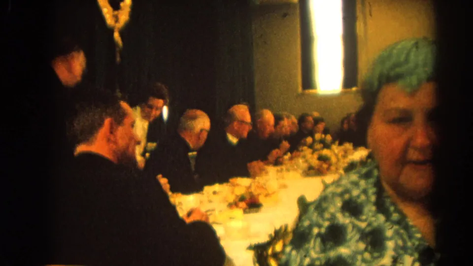 1964 Harvard, Illinois 8mm frame showing a woman in a blue patterned dress near the lens with a formal seated meeting continuing in the background.