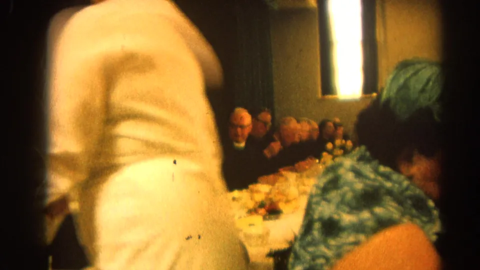 1964 Harvard, Illinois home-movie frame with a blurred person crossing the foreground and adults seated at a formal banquet table in the background.