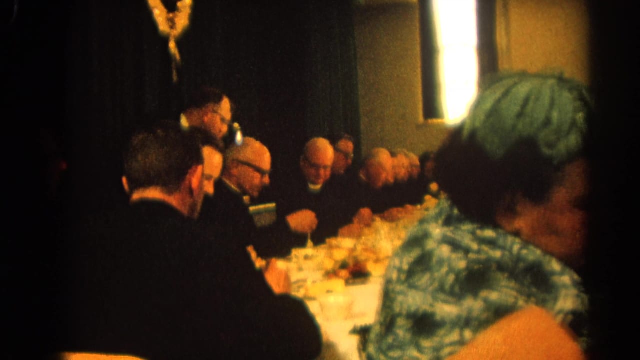 Men in black clerical clothing sit at a long dinner table as a woman in green passes in the foreground.