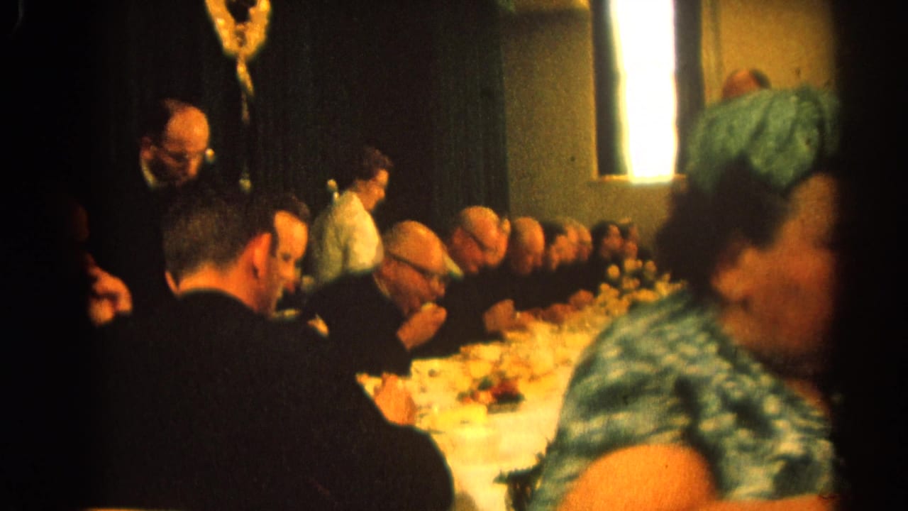 A long table of seated men in black clothing is seen behind a woman in a green patterned dress.