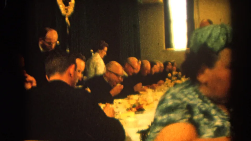 1964 Harvard, Illinois home-movie frame of men seated at a formal dinner table, with a green-dressed woman moving across the foreground.