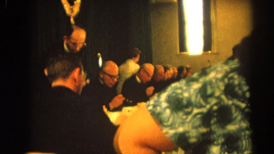1964 Harvard, Illinois 8mm frame with a woman in green patterned dress close to camera and men in black clerical clothing eating behind her.