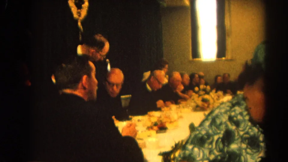 1964 Harvard, Illinois frame showing a formal dinner table with men in clerical black, plates of food, and a woman in green at the edge of the shot.
