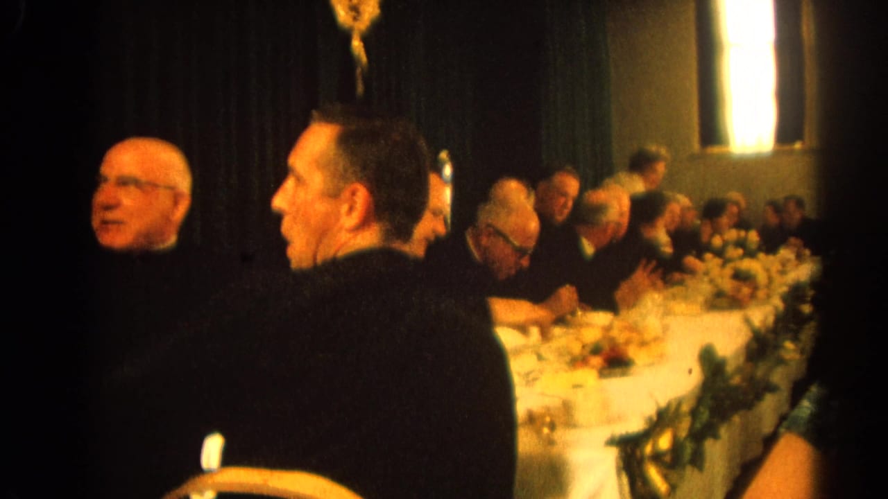 Men in dark suits and clerical attire sit at a long banquet table.