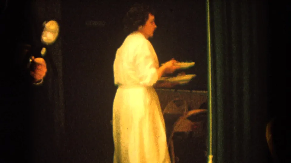 A woman in a white serving uniform carries plates near the formal dinner table in this 1964 Harvard, Illinois footage.