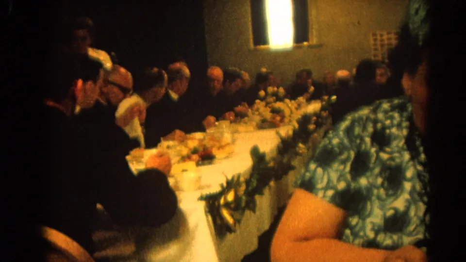 The 1964 Harvard, Illinois home movie frames diners at a clergy or family banquet, partly blocked by a woman in a blue patterned dress.