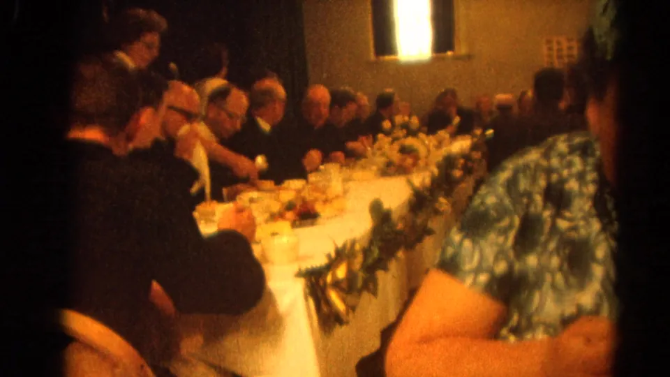 In the closing frame, clergy and guests continue dining at the long table in the 1964 Harvard, Illinois celebration footage.