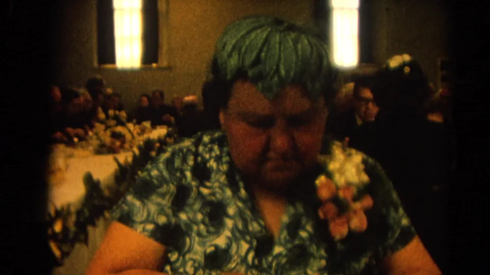 Adult woman in a green patterned dress with a floral corsage sits at a banquet table in the 1964 Harvard, Illinois home-movie footage.