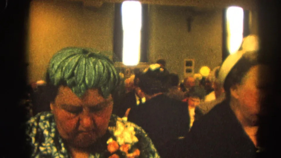 Woman in green patterned dress and corsage sits among adult banquet guests in the 1964 Harvard, Illinois home-movie scene.