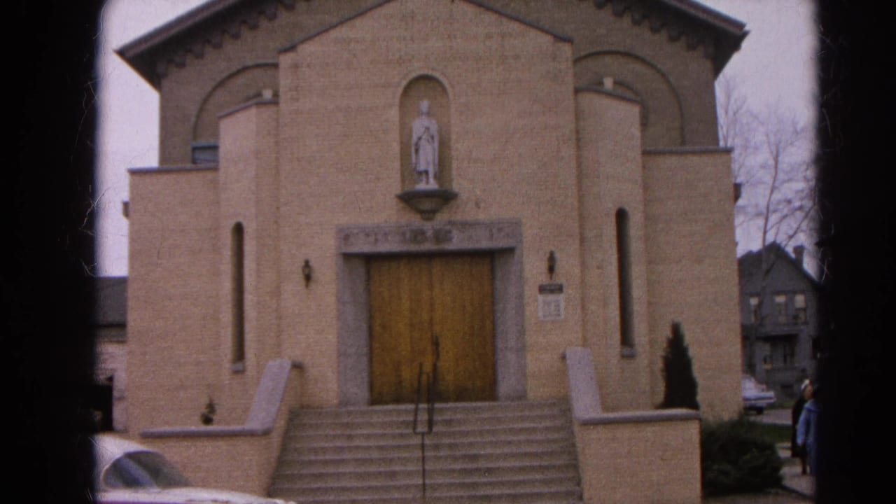 Tan brick church facade with wooden doors, steps, sidewalk, and part of a parked car.