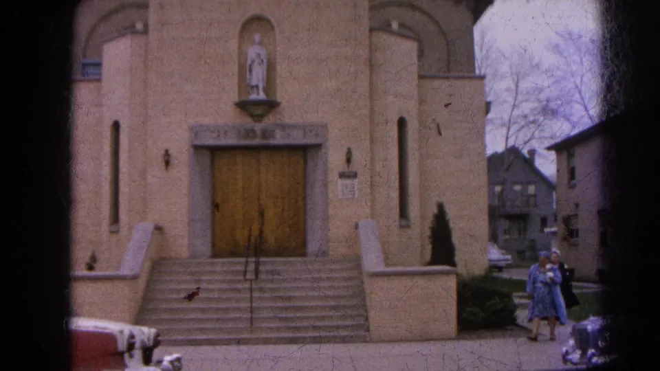 1964 Harvard, Illinois frame showing two women in long coats walking past a tan brick church with cars nearby.