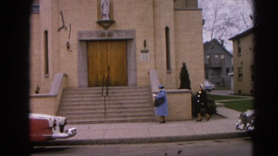 1964 8mm frame of two women in blue and dark coats walking near the church steps in Harvard, Illinois.