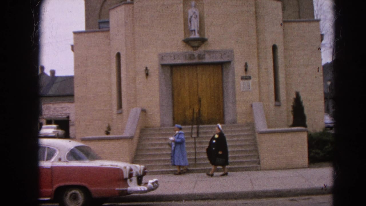 Two nuns near the steps of a beige brick church with a red-and-white sedan parked in front.