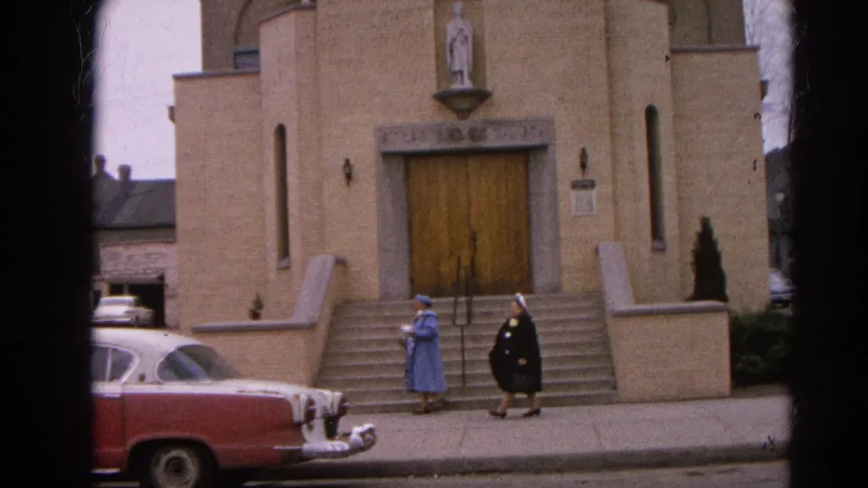 1964 Harvard, Illinois home-movie frame showing two nuns outside a beige brick church while a red-and-white sedan sits at the curb.