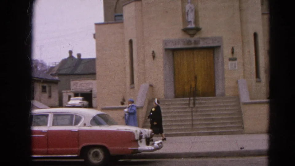 Two nuns walk past the front steps of a Harvard, Illinois church in 1964, with a mid-century red-and-white sedan at the curb.