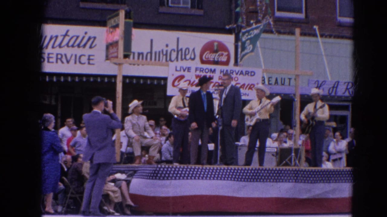 1964 Harvard, Illinois frame showing a country band on a bunting-draped stage while a cameraman films near storefront signs.