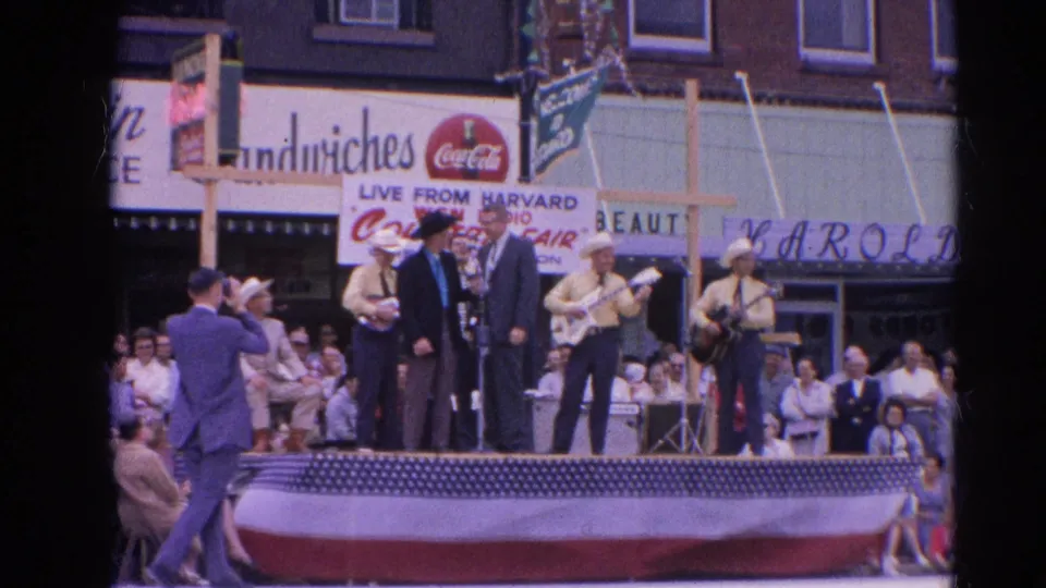 1964 Harvard, Illinois frame with a cameraman facing a stage where cowboy-hatted musicians and a suited speaker stand before spectators.