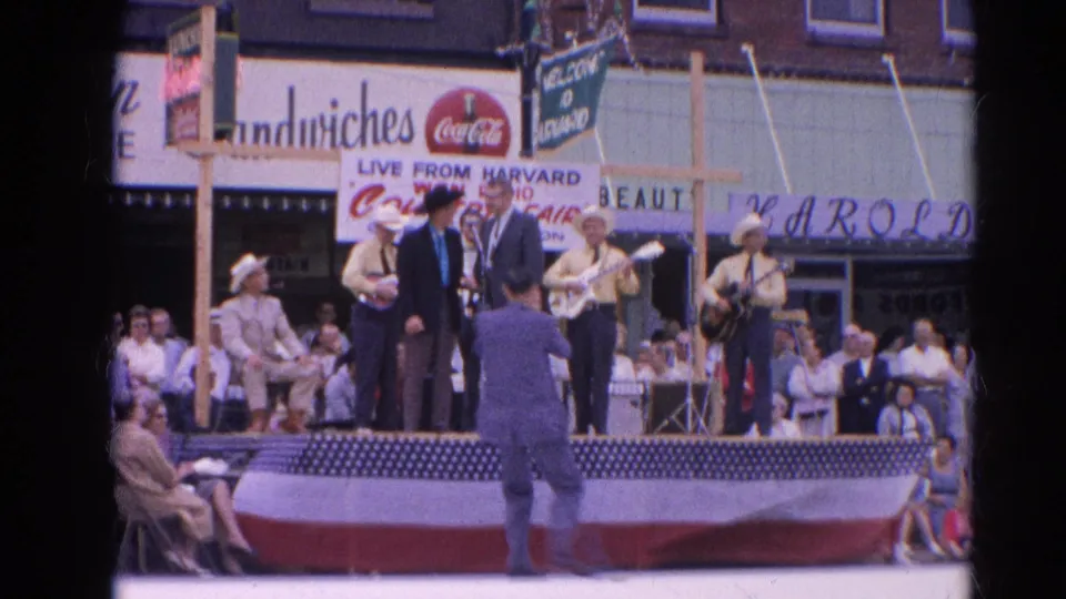 1964 Harvard, Illinois frame showing a live country performance on a decorated outdoor stage with storefront signage behind it.
