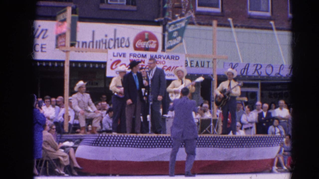 Country band and suited speakers on a flag-draped outdoor stage before storefront signs.