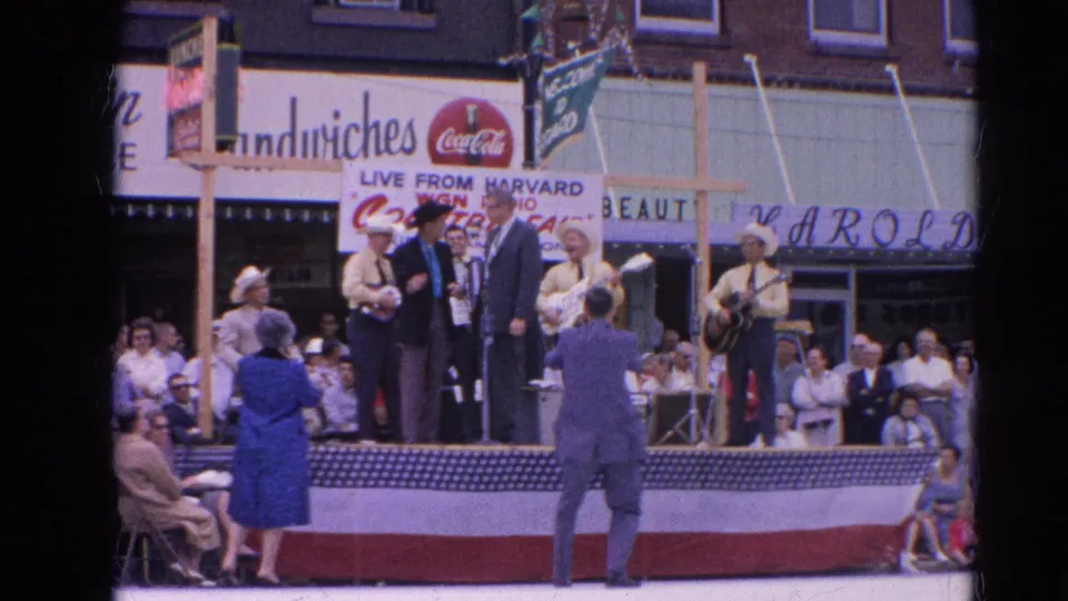 A 1964 home-movie frame shows country musicians in cowboy hats and yellow shirts beside suited speakers on a flag-draped stage in Harvard, Illinois.