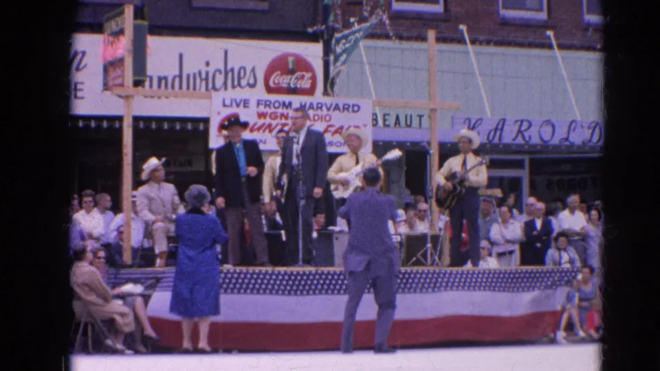 The frame captures a small-town Illinois country music event with musicians, suited speakers, spectators, and commercial signs behind the flag-draped stage.