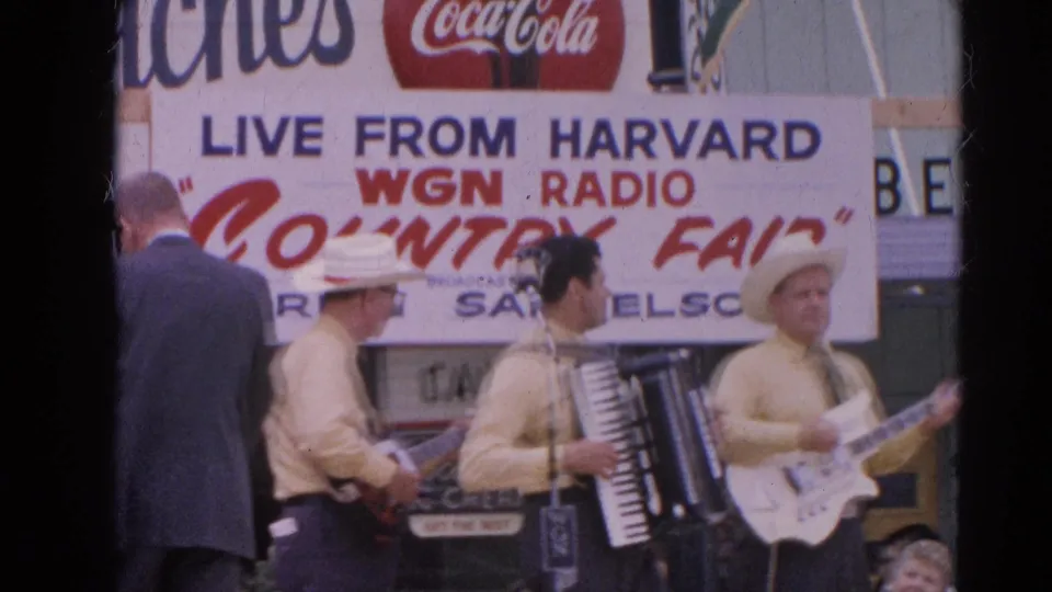 The frame shows the Harvard WGN Radio Country Fair banner above musicians and a man in a dark suit near the stage.