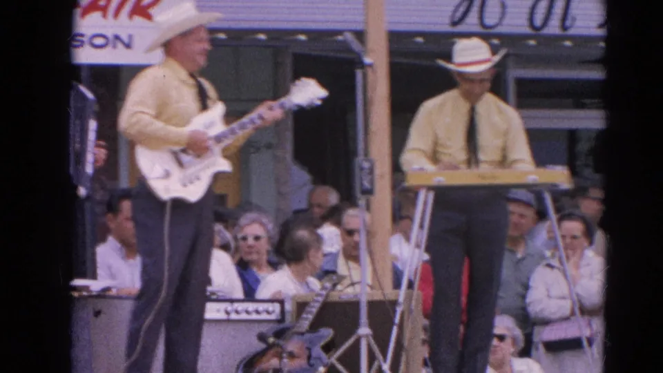 The country fair band continues playing on the outdoor stage, with guitar, keyboard, microphone stands, and a watching crowd visible.