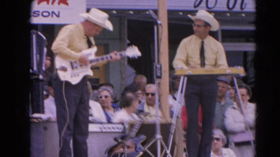 1964 Harvard, Illinois home-movie frame showing two musicians in matching shirts and white cowboy hats playing guitar and keyboard outdoors.