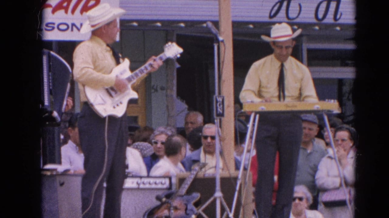 Home-movie frame from Harvard, Illinois in 1964 showing a guitarist in a white hat facing a keyboard player on an outdoor stage.