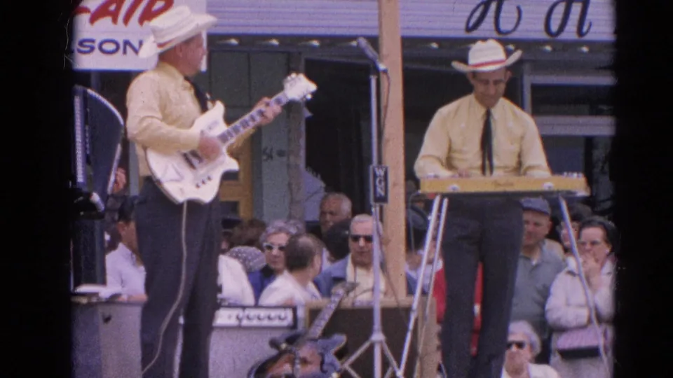 Home-movie frame from Harvard, Illinois in 1964 showing a guitarist in a white hat facing a keyboard player on an outdoor stage.