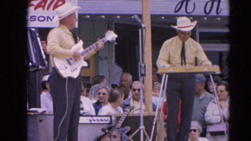 1964 Harvard, Illinois 8mm frame showing two matching-shirt musicians performing guitar and keyboard before an outdoor crowd.