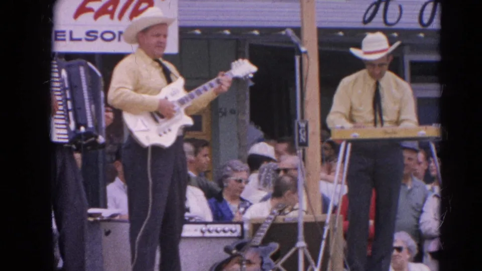 Final 1964 Harvard, Illinois home-movie frame of an outdoor musical act with guitar, keyboard, cowboy hats, and a watching crowd.