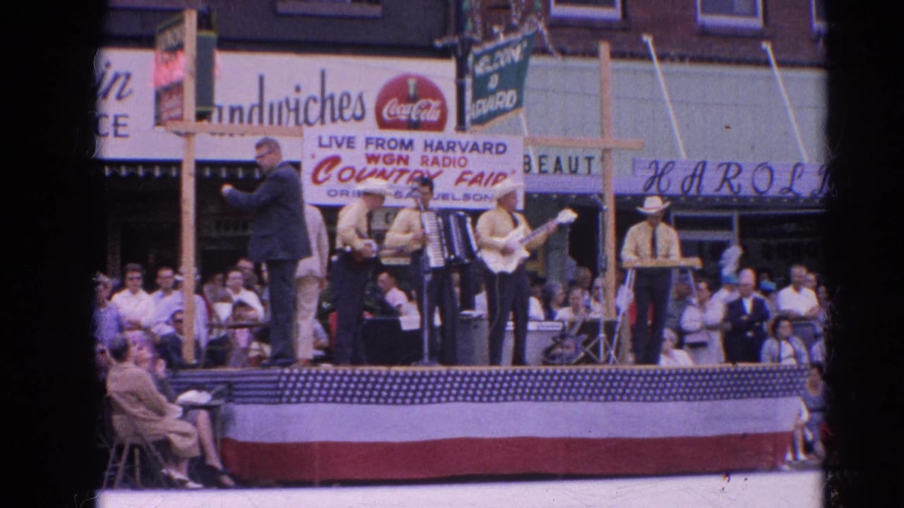 At the Harvard Country Fair in 1964, a suited announcer faces the country band on a decorated street stage with storefront signs behind them.