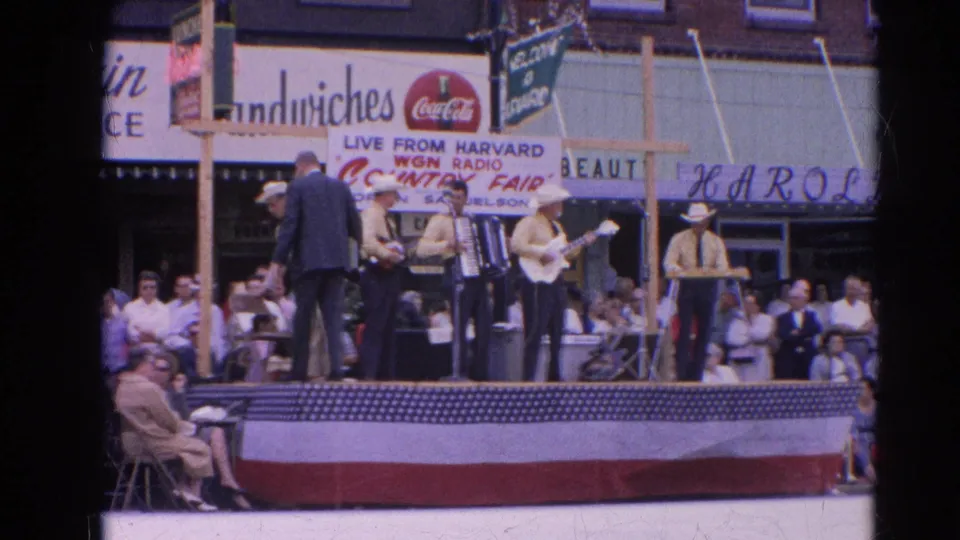 The 1964 Harvard, Illinois fair stage shows a suited host speaking or conducting near musicians as a street crowd watches.