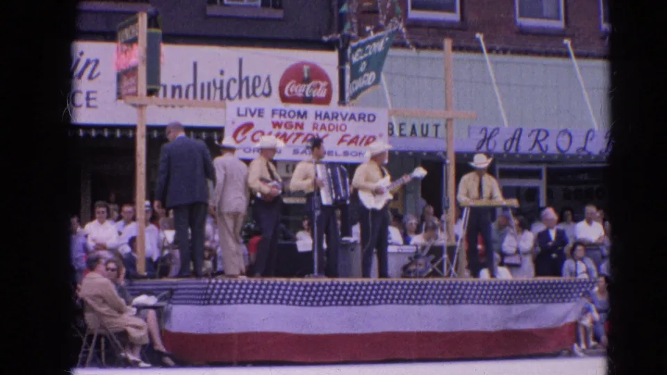 Two suited men stand with the country musicians on the Harvard, Illinois fair stage, with WGN Radio and Coca-Cola signage visible behind them.
