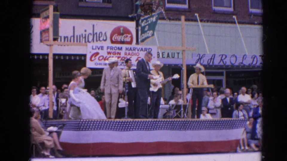 A 1964 Harvard, Illinois public performance shows dancers, musicians, an accordion, guitar, bunting, and spectators in front of storefronts.