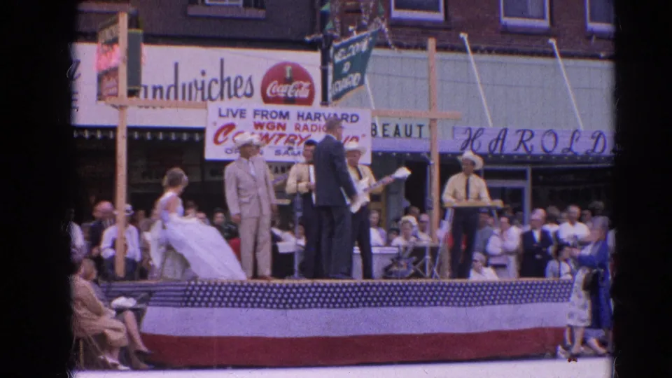Street-stage country music performance in Harvard, Illinois, 1964, with a dancing couple, musicians, bunting, and a gathered sidewalk crowd.