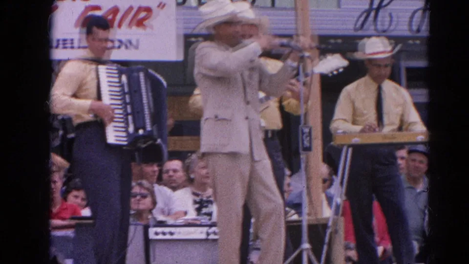 1964 Harvard, Illinois fair stage with a violinist in a light suit, accordion player, keyboard musician, and watching crowd.