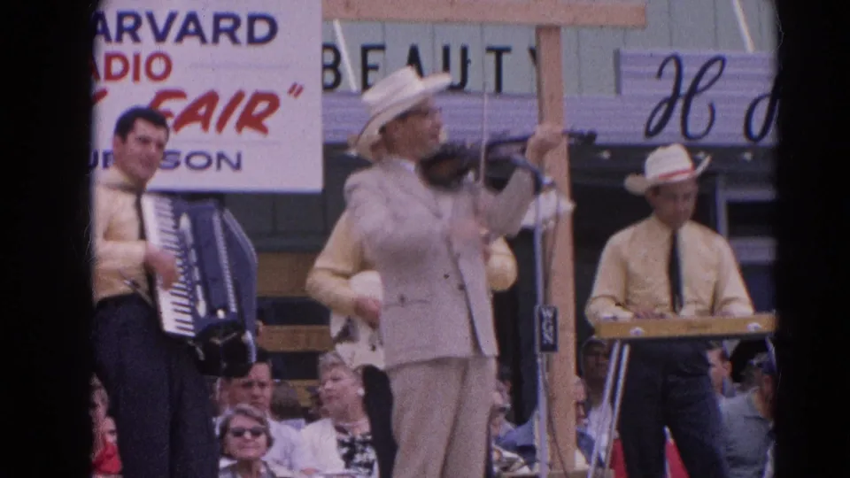 1964 home-movie frame of a Harvard, Illinois fair band with violin, accordion, keyboard, hats, signage, and spectators.