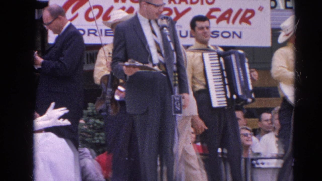 At the Harvard, Illinois county fair, a suited announcer stands at microphones while an accordion player and other musicians wait nearby.