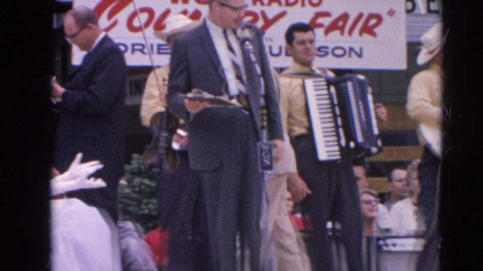 At the Harvard, Illinois county fair, a suited announcer stands at microphones while an accordion player and other musicians wait nearby.