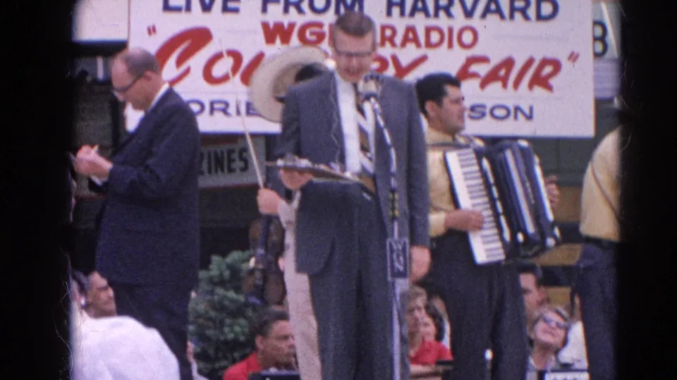 A violinist and suited announcer stand near microphones with an accordion player behind them at the 1964 Harvard, Illinois fair performance.